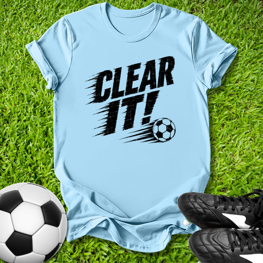Light blue t-shirt with 'CLEAR IT!' text and soccer ball graphic on grass with soccer balls and shoes.