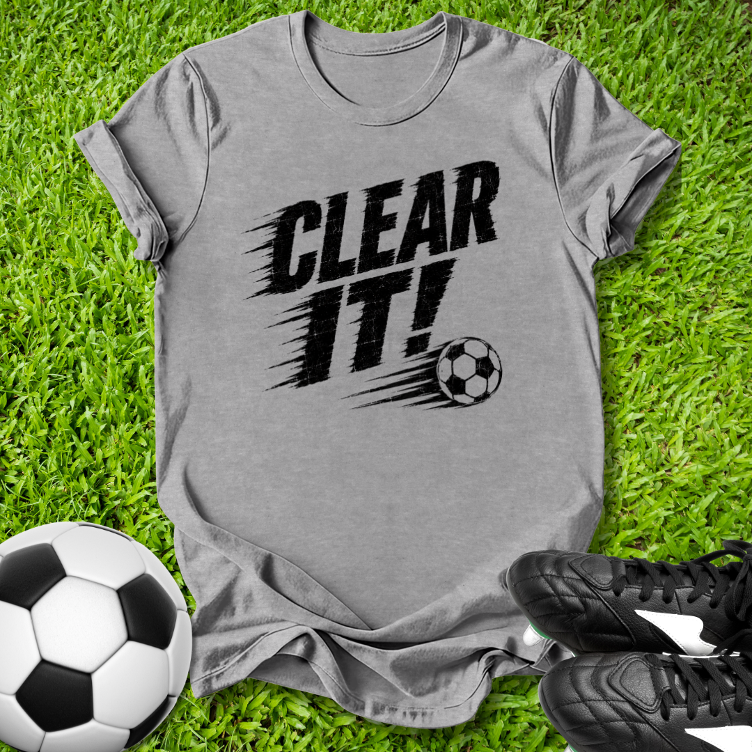 Gray t-shirt with 'CLEAR IT!' text and soccer ball graphic on grass with soccer balls and shoes.