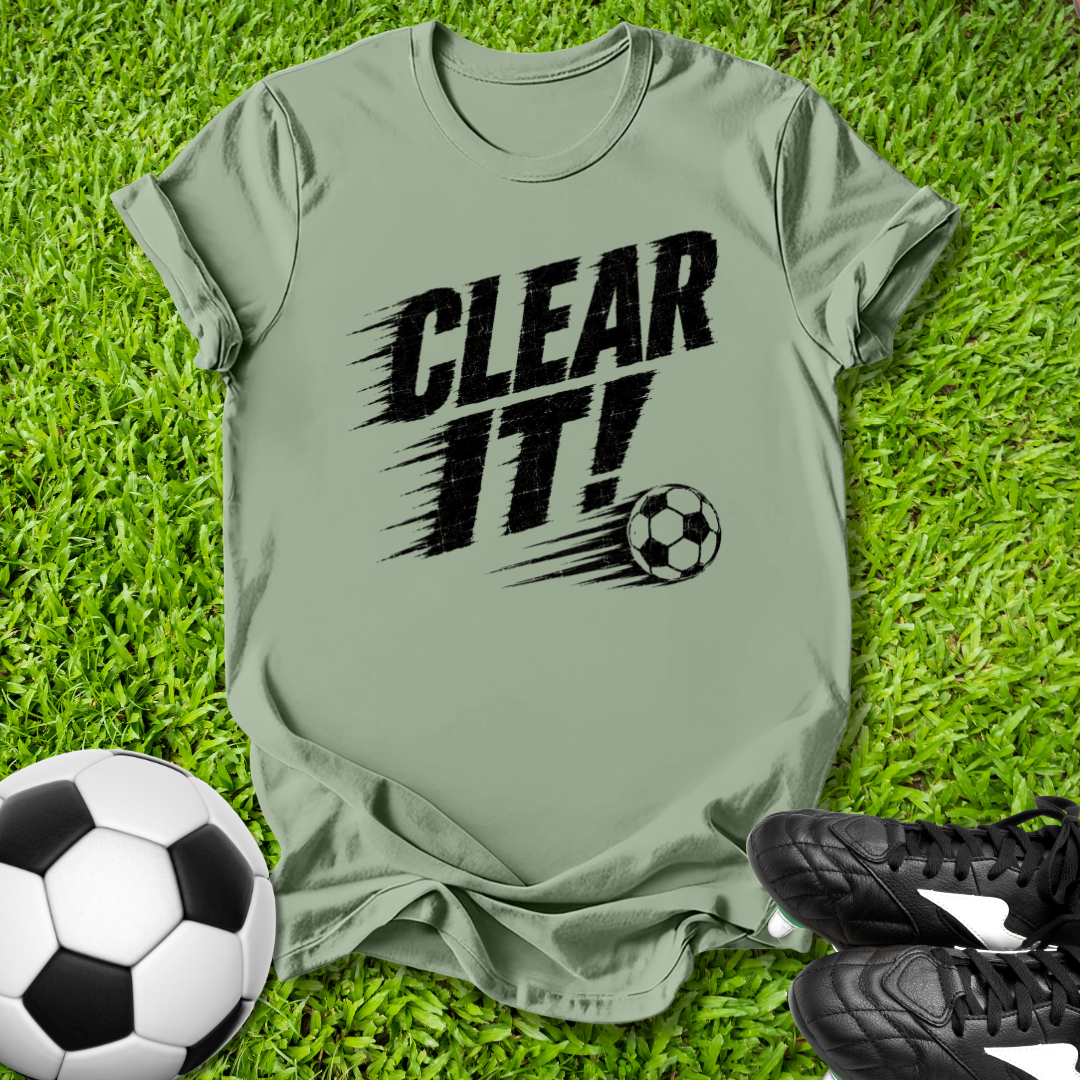 Green t-shirt with 'CLEAR IT!' text and soccer ball graphic on grass with soccer ball and shoes.