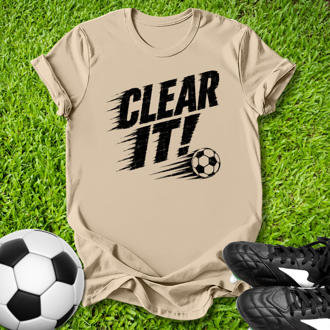 Beige t-shirt with 'CLEAR IT!' text and soccer ball graphic on grass with soccer ball and shoes.