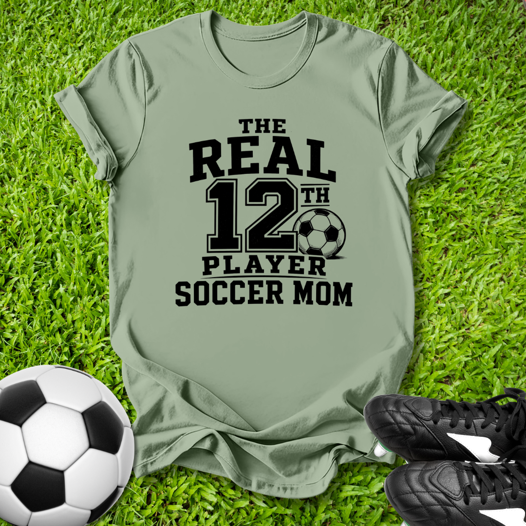 Green t-shirt with 'The Real 12th Player Soccer Mom' text on grass with soccer ball and shoes.