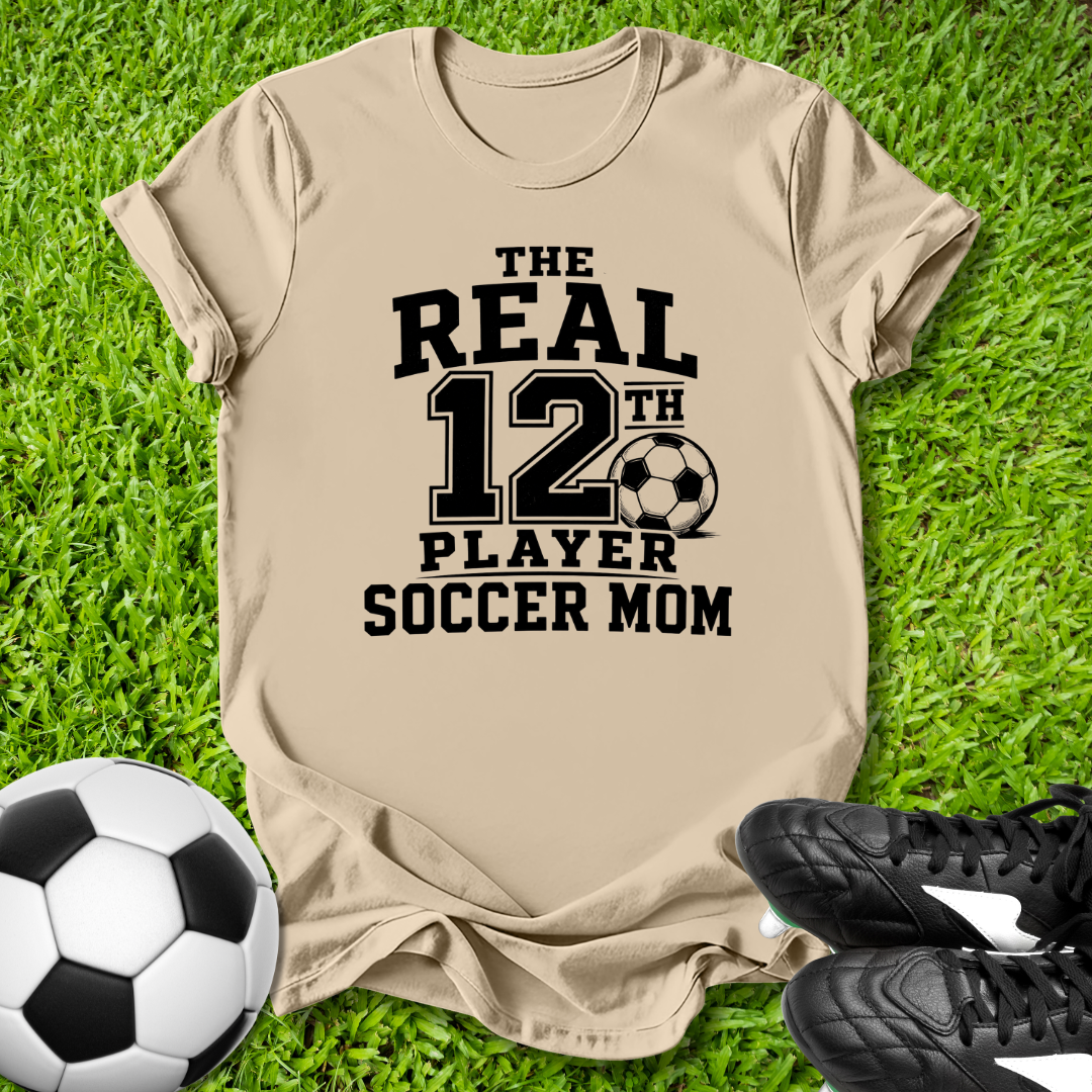 Beige t-shirt with 'The Real 12th Player Soccer Mom' text on grass with soccer ball and shoes.