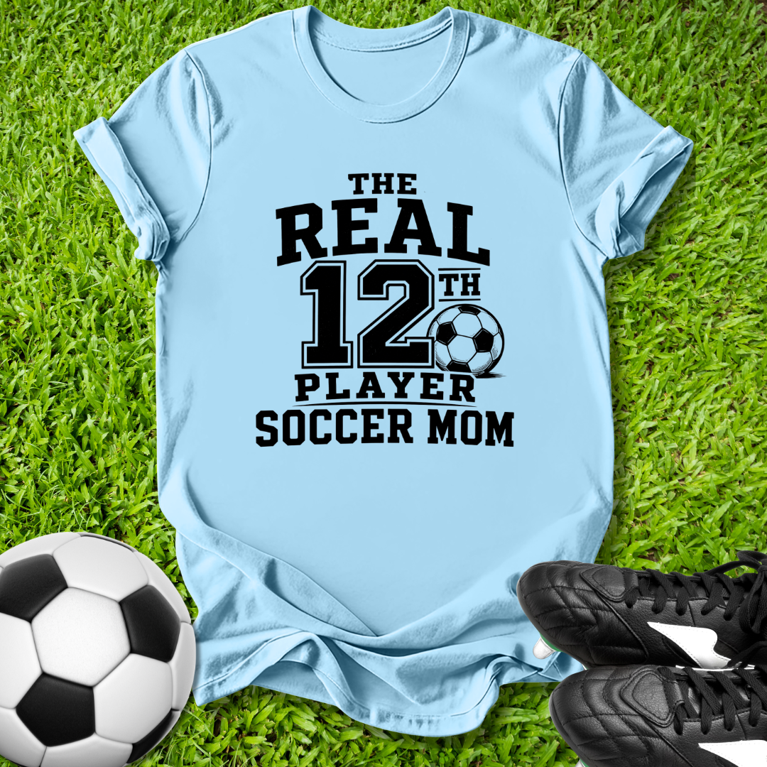 Light blue t-shirt with 'The Real 12th Player Soccer Mom' text on grass with soccer ball and shoes.
