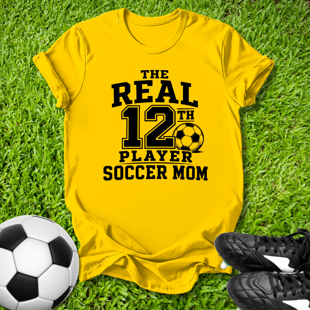 Yellow t-shirt with 'The Real 12th Player Soccer Mom' text on grass with soccer ball and shoes.