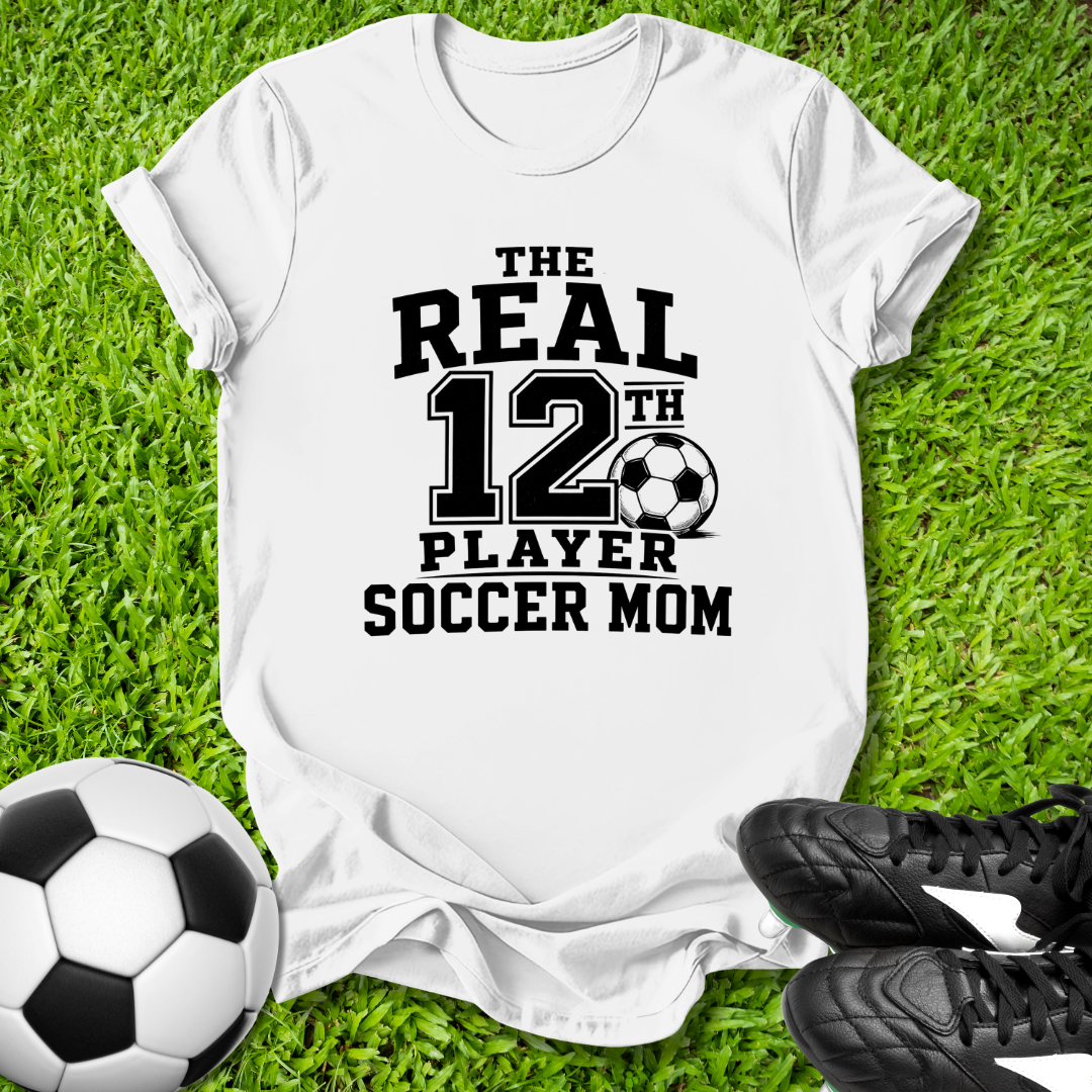 White t-shirt with 'The Real 12th Player Soccer Mom' text on grass with soccer ball and shoes.