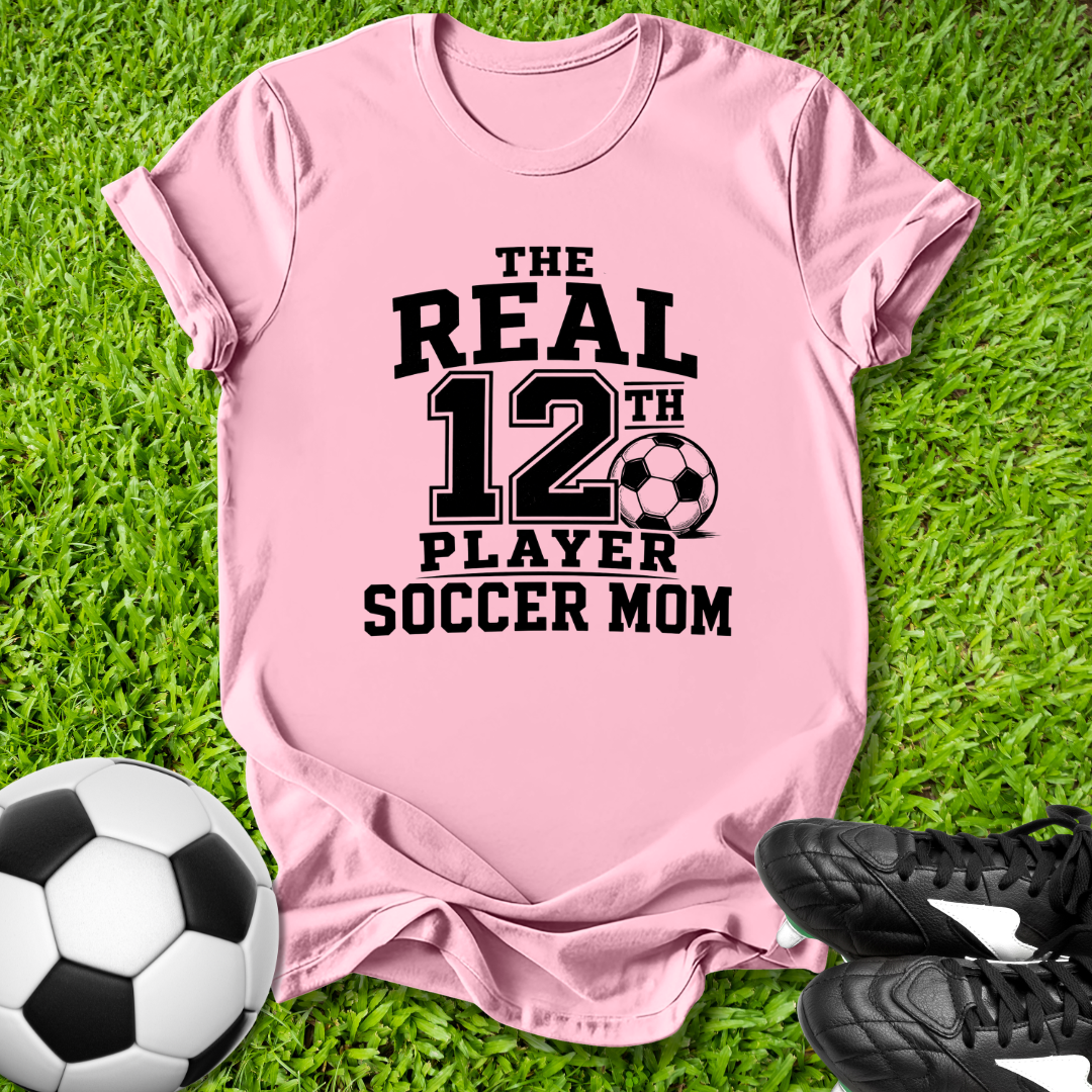 Pink t-shirt with 'The Real 12th Player Soccer Mom' text on grass with soccer ball and shoes.