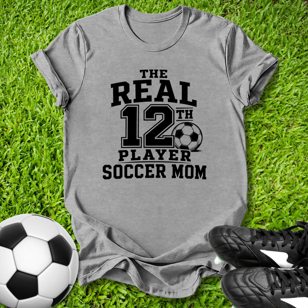 Gray t-shirt with 'The Real 12th Player Soccer Mom' text on grass with soccer ball and shoes.