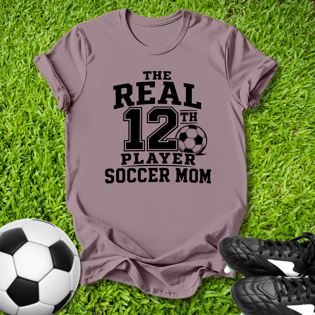 Purple t-shirt with 'The Real 12th Player Soccer Mom' text on grass with soccer ball and shoes.