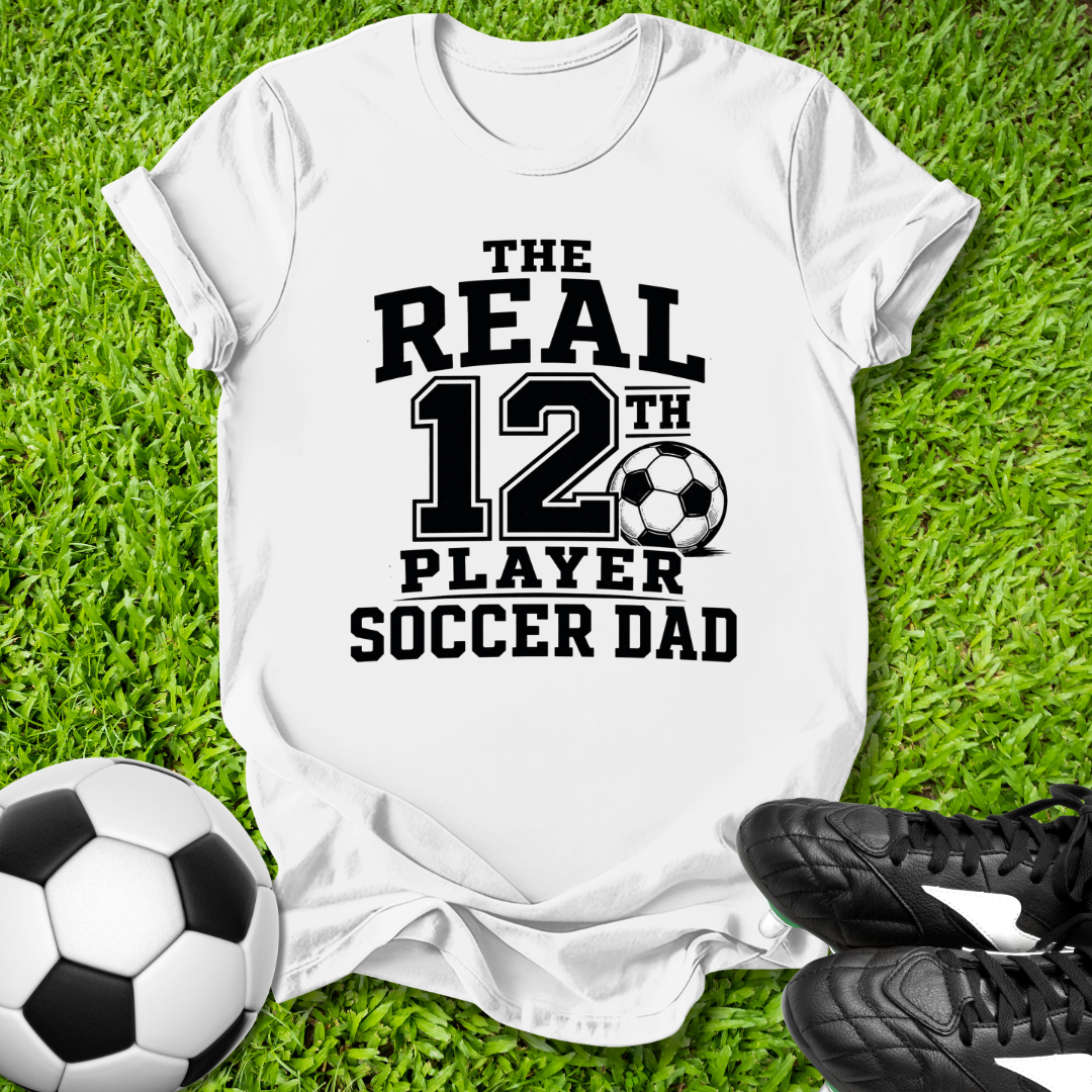 White t-shirt with 'The Real 12th Player Soccer Dad' text on grass with soccer ball and shoes.