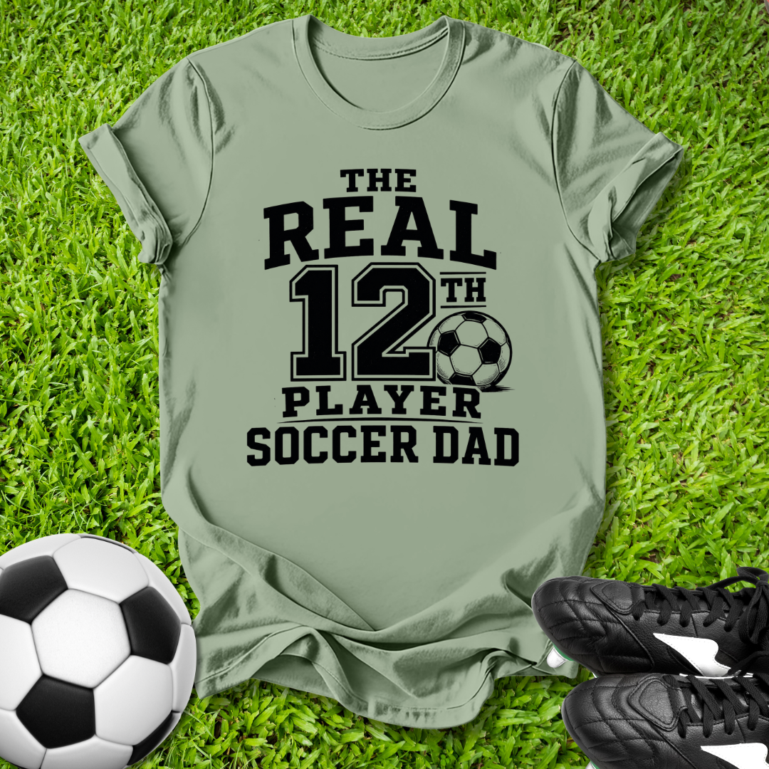 Green t-shirt with 'The Real 12th Player Soccer Dad' text on grass with soccer ball and shoes.
