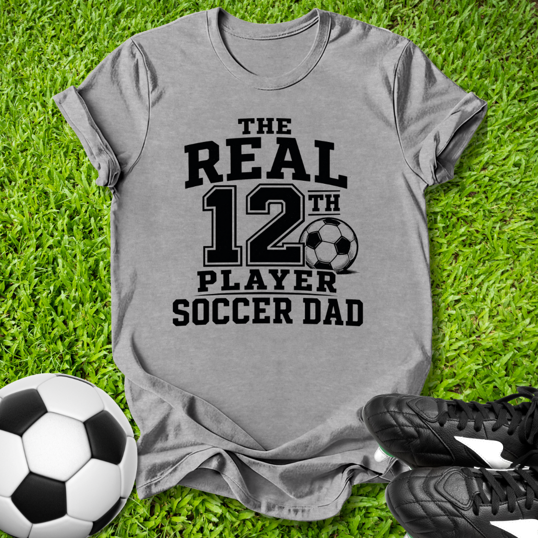 Gray t-shirt with 'The Real 12th Player Soccer Dad' text on grass with soccer ball and shoes.
