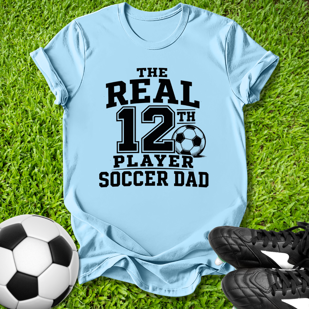 Light blue t-shirt with 'The Real 12th Player Soccer Dad' text on grass with soccer ball and shoes.