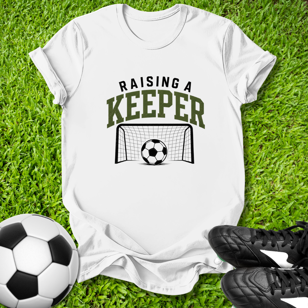 White t-shirt with 'Raising a Keeper' graphic on grass with soccer ball and cleats.