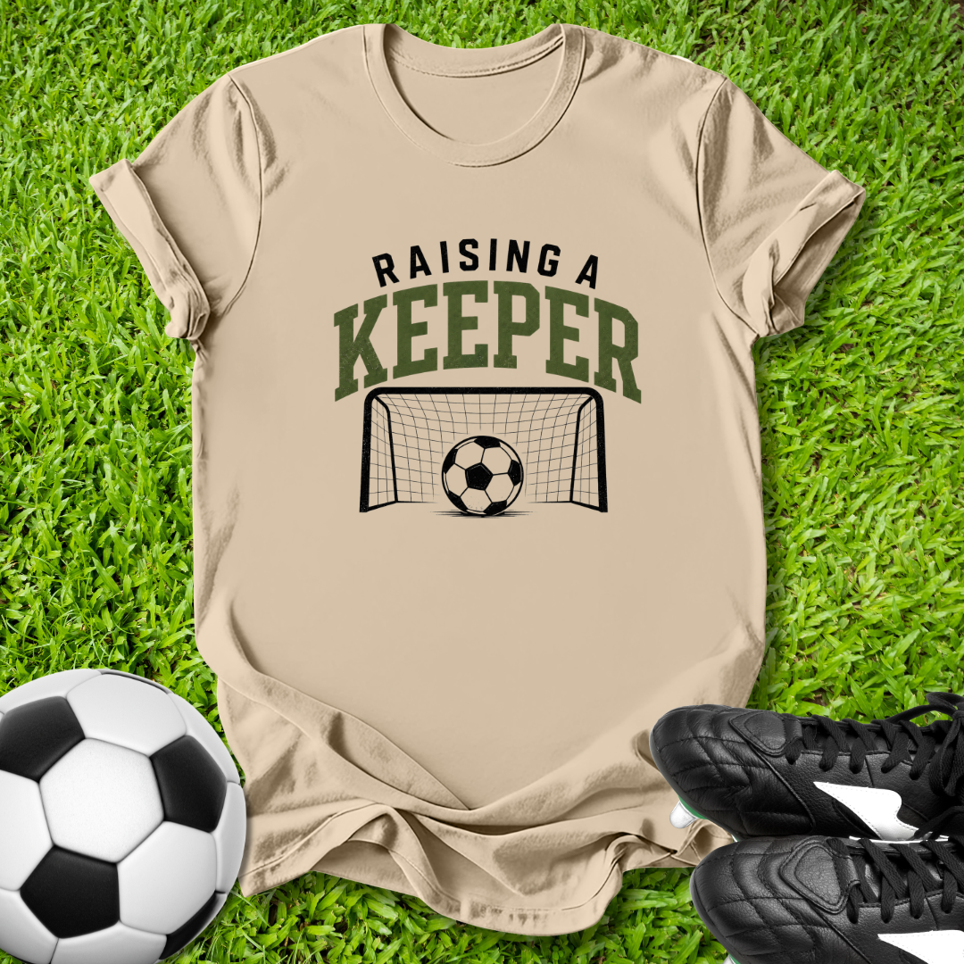 Beige t-shirt with 'Raising a Keeper' graphic on grass with soccer ball and shoes.
