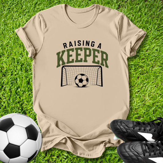 Beige t-shirt with 'Raising a Keeper' graphic on grass with soccer ball and shoes.