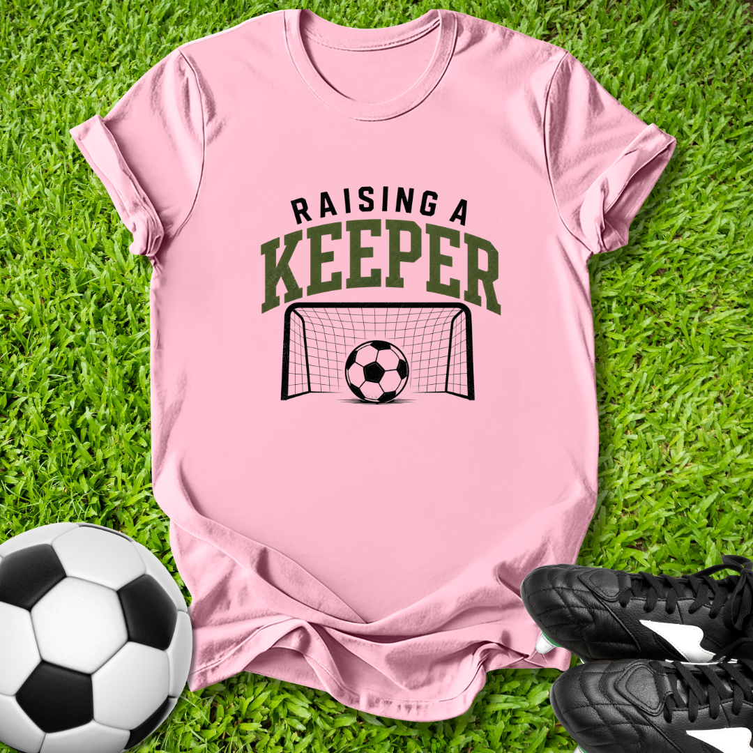 Pink t-shirt with 'Raising a Keeper' text and soccer graphics on grass with soccer ball and shoes.