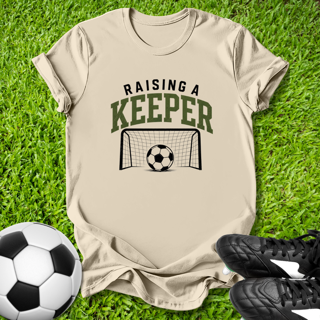 Beige t-shirt with 'Raising a Keeper' graphic on grass with soccer balls and shoes.