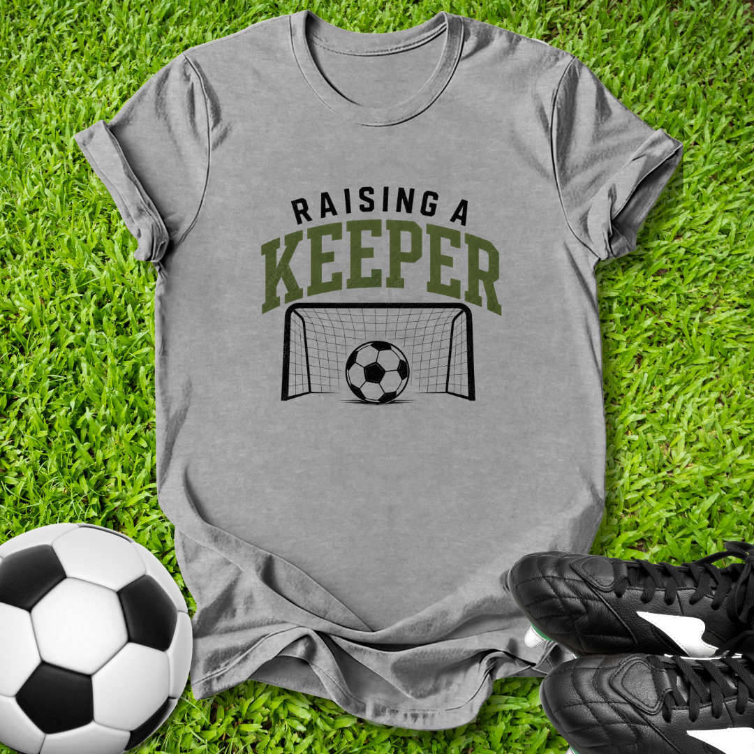 Gray t-shirt with 'Raising a Keeper' text and soccer graphics on grass with soccer balls and shoes.