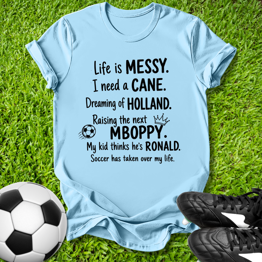 Light blue t-shirt with humorous text on grass with soccer ball and shoes
