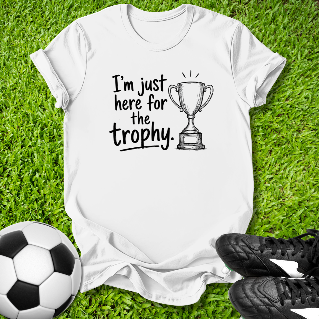 White t-shirt with 'I'm just here for the trophy' text and soccer ball on grass