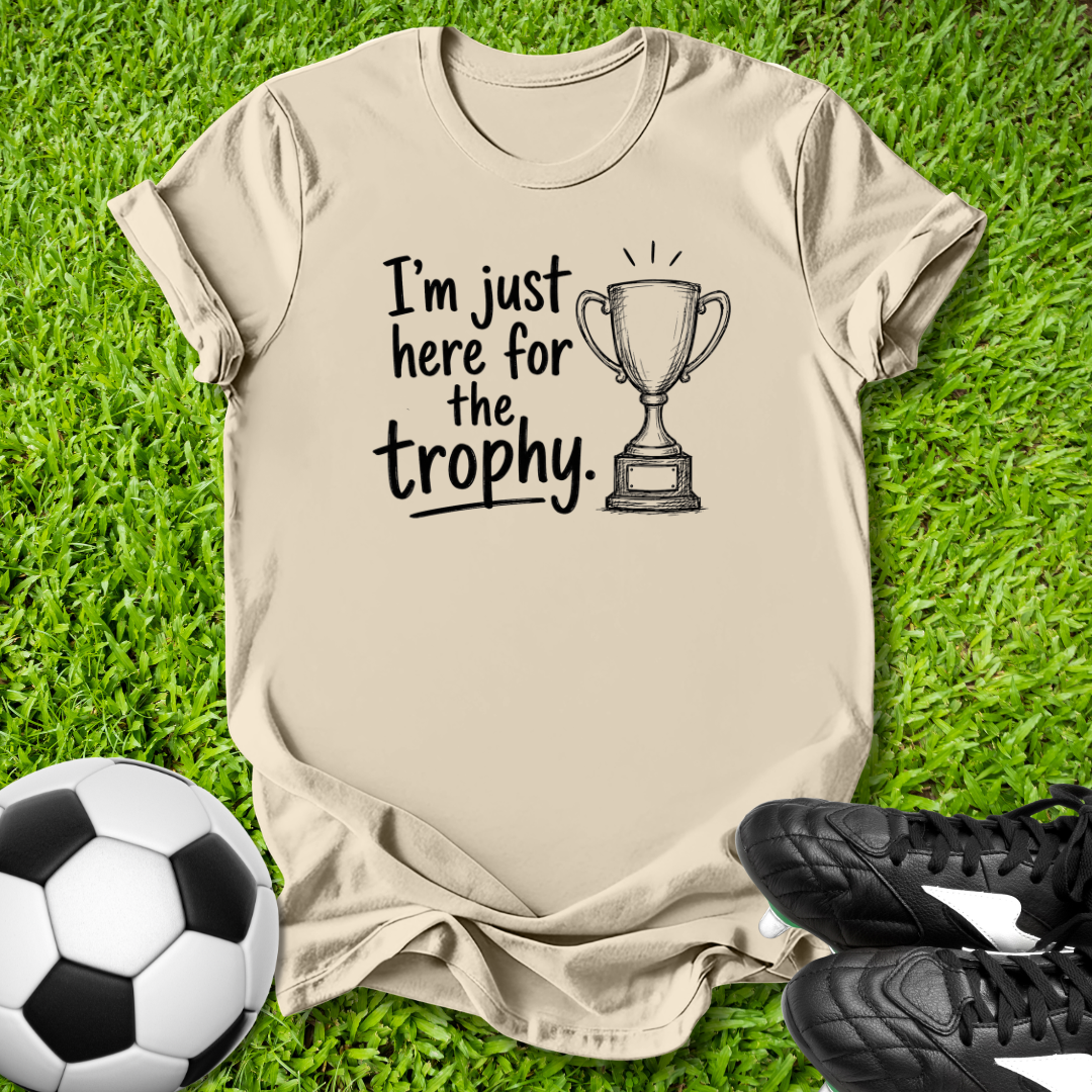 Beige t-shirt with 'I'm just here for the trophy' text and trophy graphic on grass with soccer ball and shoes.