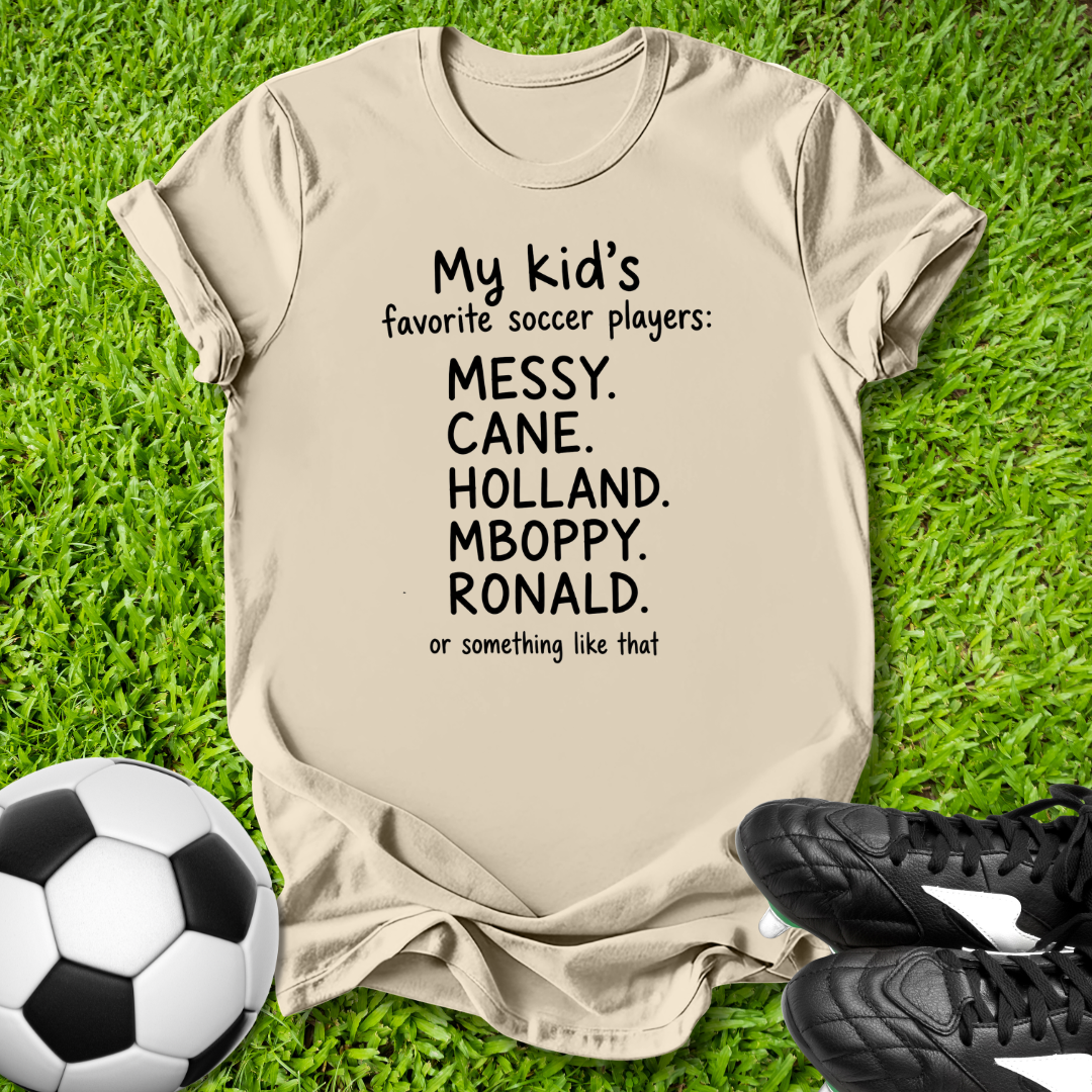 Beige t-shirt with humorous soccer player names on grass with soccer ball and shoes.