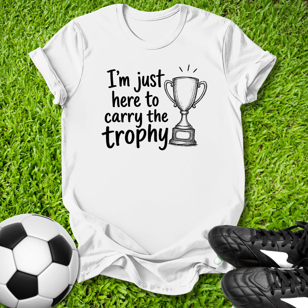 White t-shirt with 'I'm just here to carry the trophy' text and soccer ball and shoes on grass