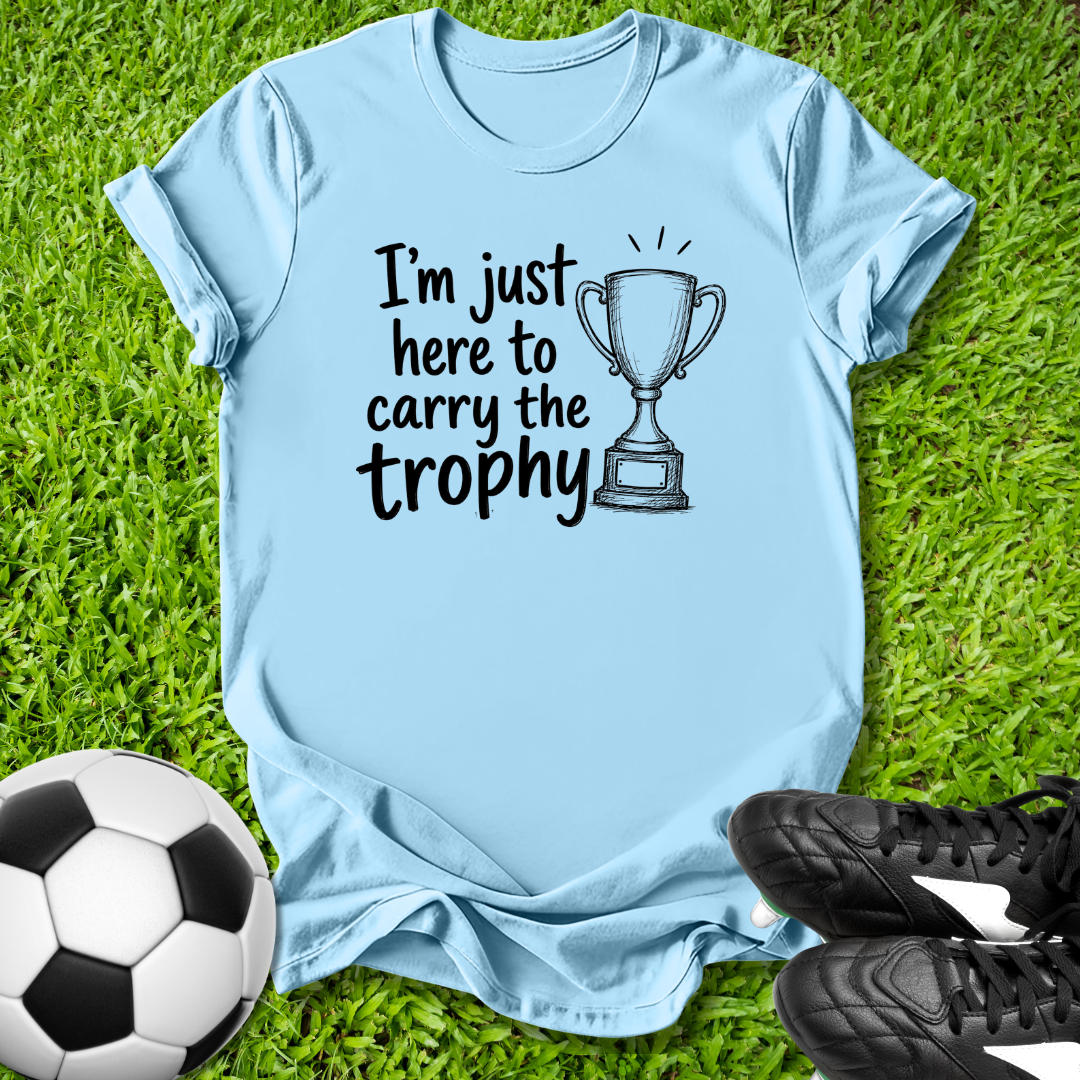 Light blue t-shirt with 'I'm just here to carry the trophy' text and soccer ball and shoes on grass