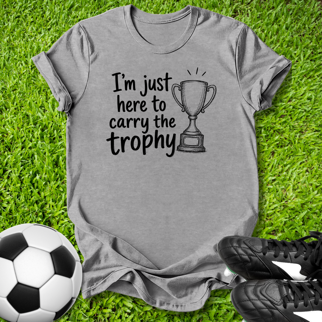 Gray t-shirt with 'I'm just here to carry the trophy' text and soccer ball and shoes on grass