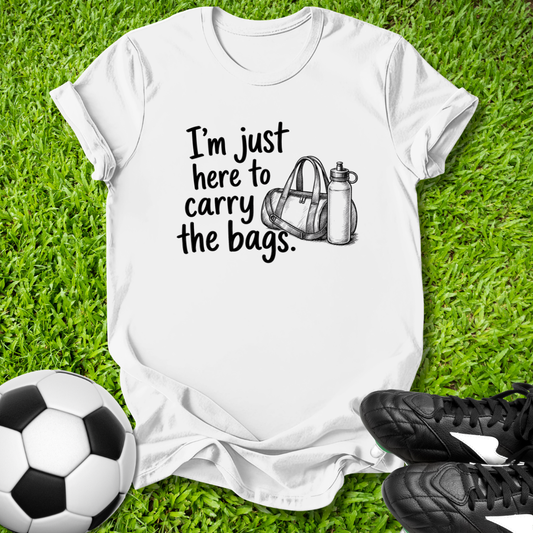 White t-shirt with 'I'm just here to carry the bags' text and graphic on grass with soccer ball and shoes.