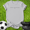 A light gray short-sleeve t-shirt with black cursive text reading 'carpal queen, snack handler, biggest fan.' and a small heart, laid flat on green grass with a soccer ball and black cleats as props.