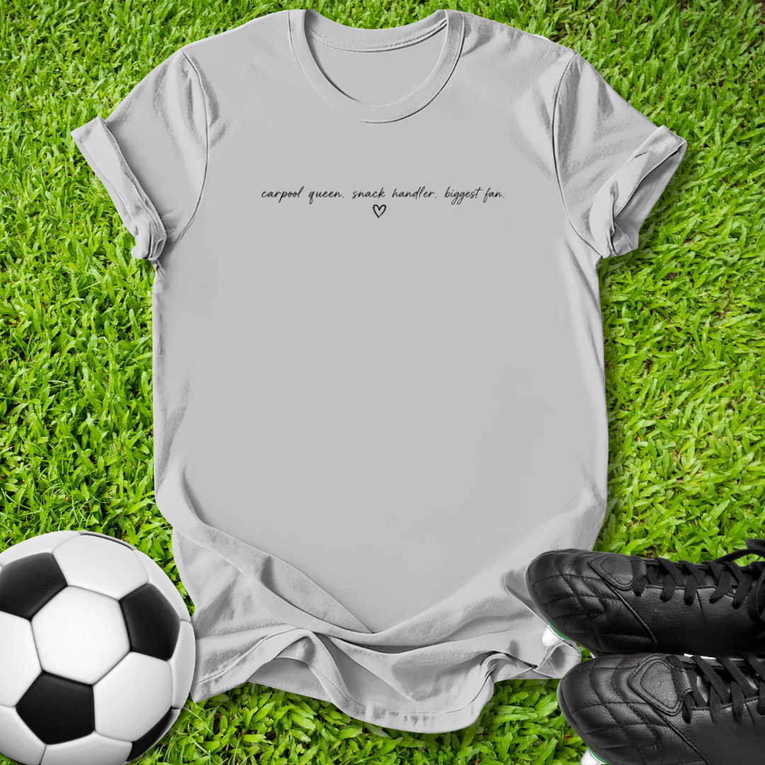 A light gray short-sleeve t-shirt with black cursive text reading 'carpal queen, snack handler, biggest fan.' and a small heart, laid flat on green grass with a soccer ball and black cleats as props.