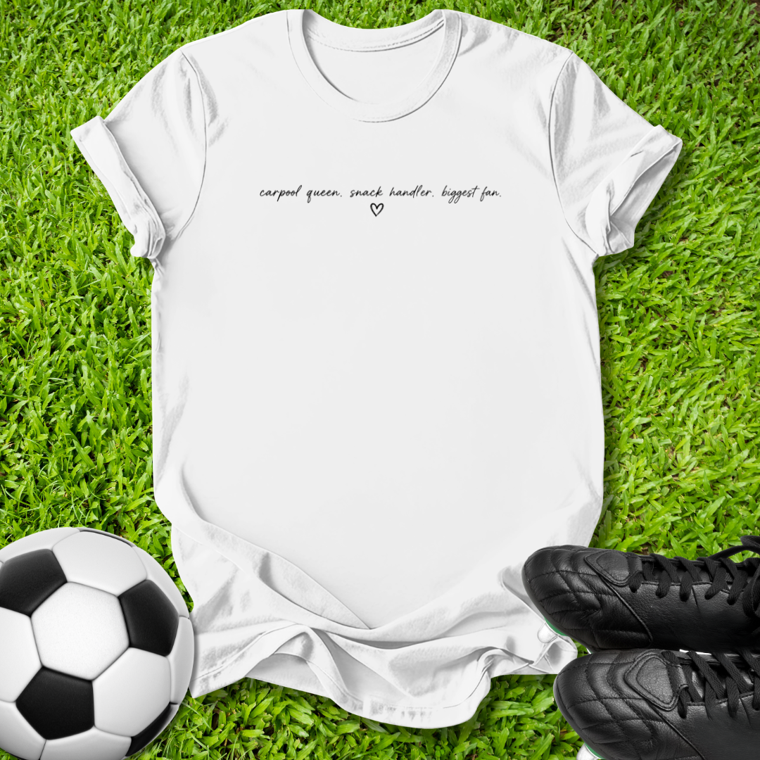 A white short-sleeve graphic T-shirt laid flat on green grass, with a small black script slogan across the chest and a tiny heart below it. A soccer ball appears at the lower left, and black cleats are at the lower right, creating a casual outdoor-style flat lay.