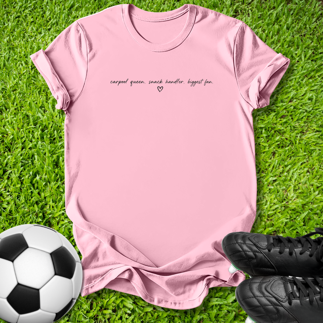 A light pink short-sleeve t-shirt with black cursive text reading 'carpal queen, snack handler biggest fan.' and a small heart, laid flat on green grass with a soccer ball and black cleats as props.