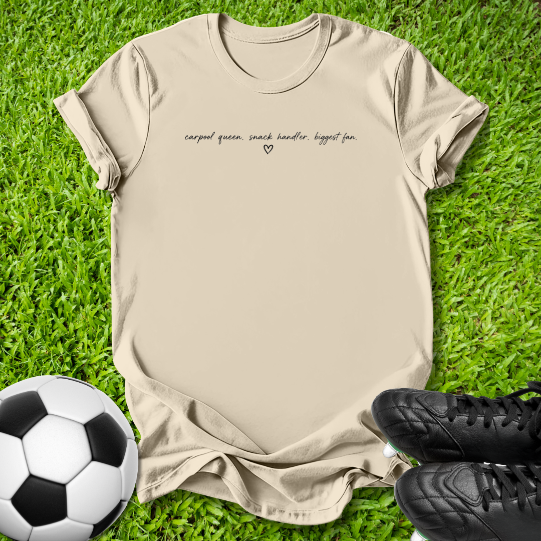 A beige graphic t-shirt laid flat on green grass, styled with a soccer ball on the left and black cleats on the right. The t-shirt features the text 'carpal queen, snack handler biggest fan.