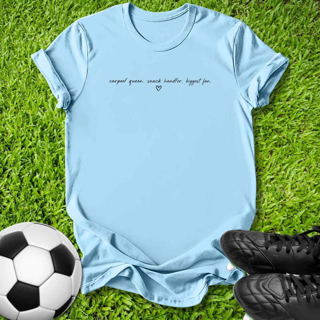 A light blue short-sleeve t-shirt with the text 'carpal queen, snack handler, biggest fan.' and a small heart printed on the chest, laid flat on green grass with a soccer ball and black cleats as props.