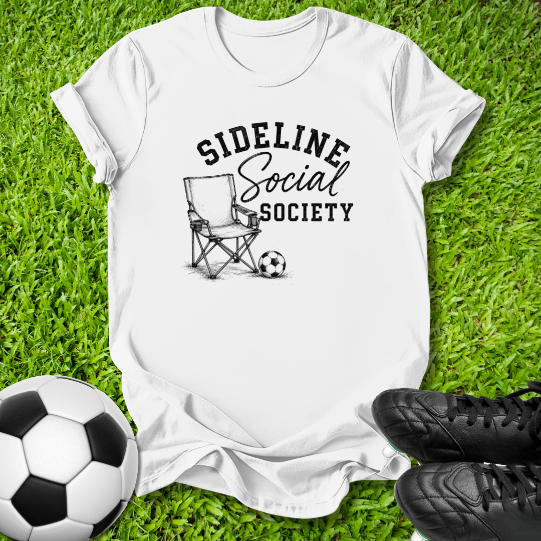 A white short-sleeve graphic T-shirt laid flat on green grass, with a black printed design reading 'SIDELINE Social SOCIETY' around a folding chair and soccer ball. A soccer ball and black cleats are placed around the shirt as styling props.