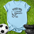 A light blue graphic t-shirt laid flat on green grass, featuring a black printed design that reads 'SIDELINE Social SOCIETY' with a folding chair and soccer ball illustration. A soccer ball and black cleats are placed around the shirt as props.
