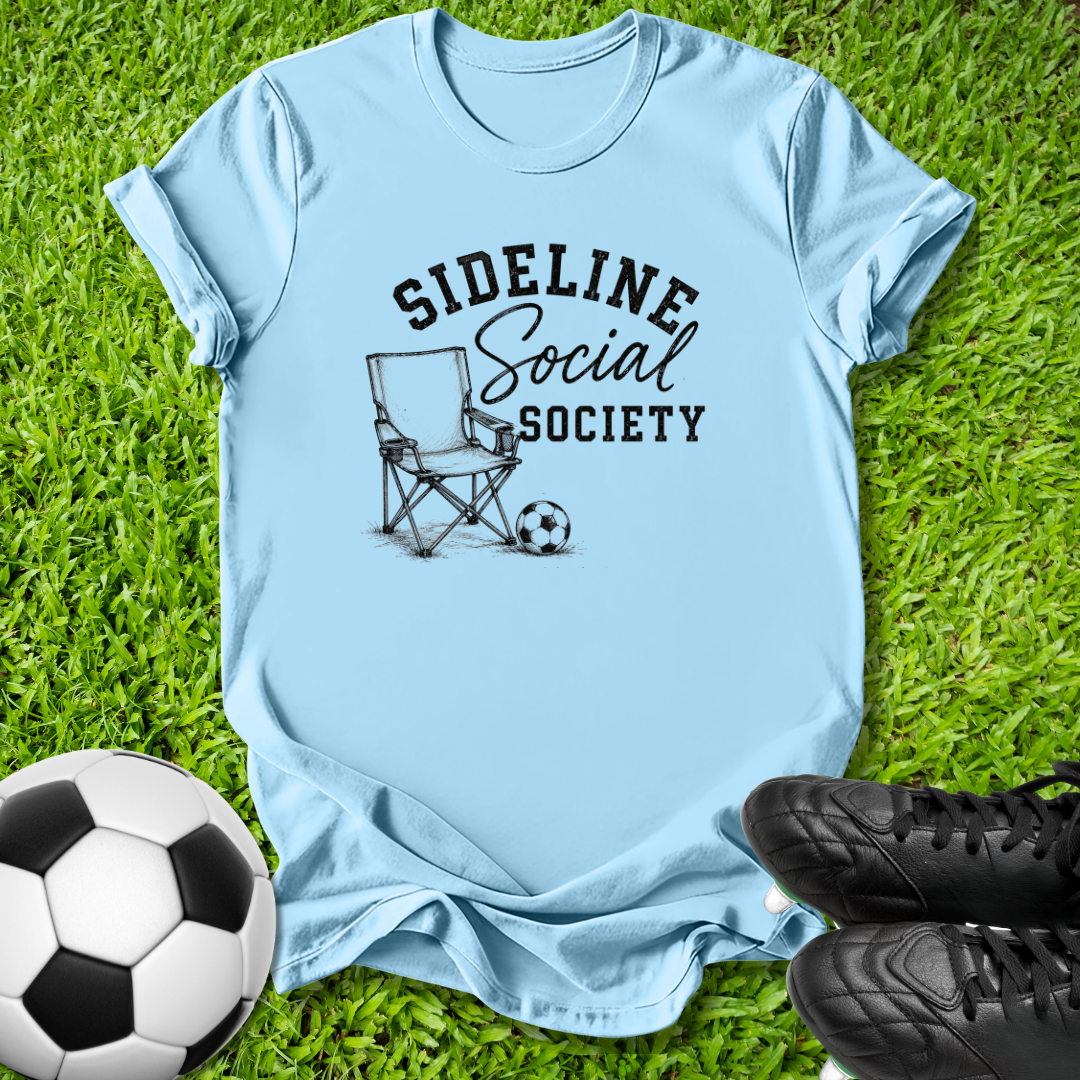 A light blue graphic t-shirt laid flat on green grass, featuring a black printed design that reads 'SIDELINE Social SOCIETY' with a folding chair and soccer ball illustration. A soccer ball and black cleats are placed around the shirt as props.
