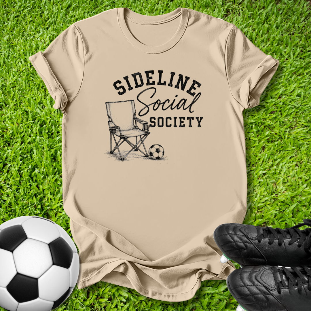 A beige graphic t-shirt laid flat on green grass, featuring a black print that reads 'SIDELINE Social SOCIETY' with a folding chair and soccer ball illustration. A soccer ball is on the left, and black cleats are on the right.