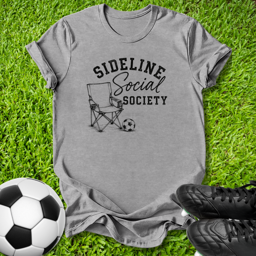 A heather gray graphic t-shirt laid flat on green grass, featuring a black print that reads 'SIDELINE Social SOCIETY' with a folding chair and soccer ball illustration. A soccer ball and black cleats are placed around the shirt as props.