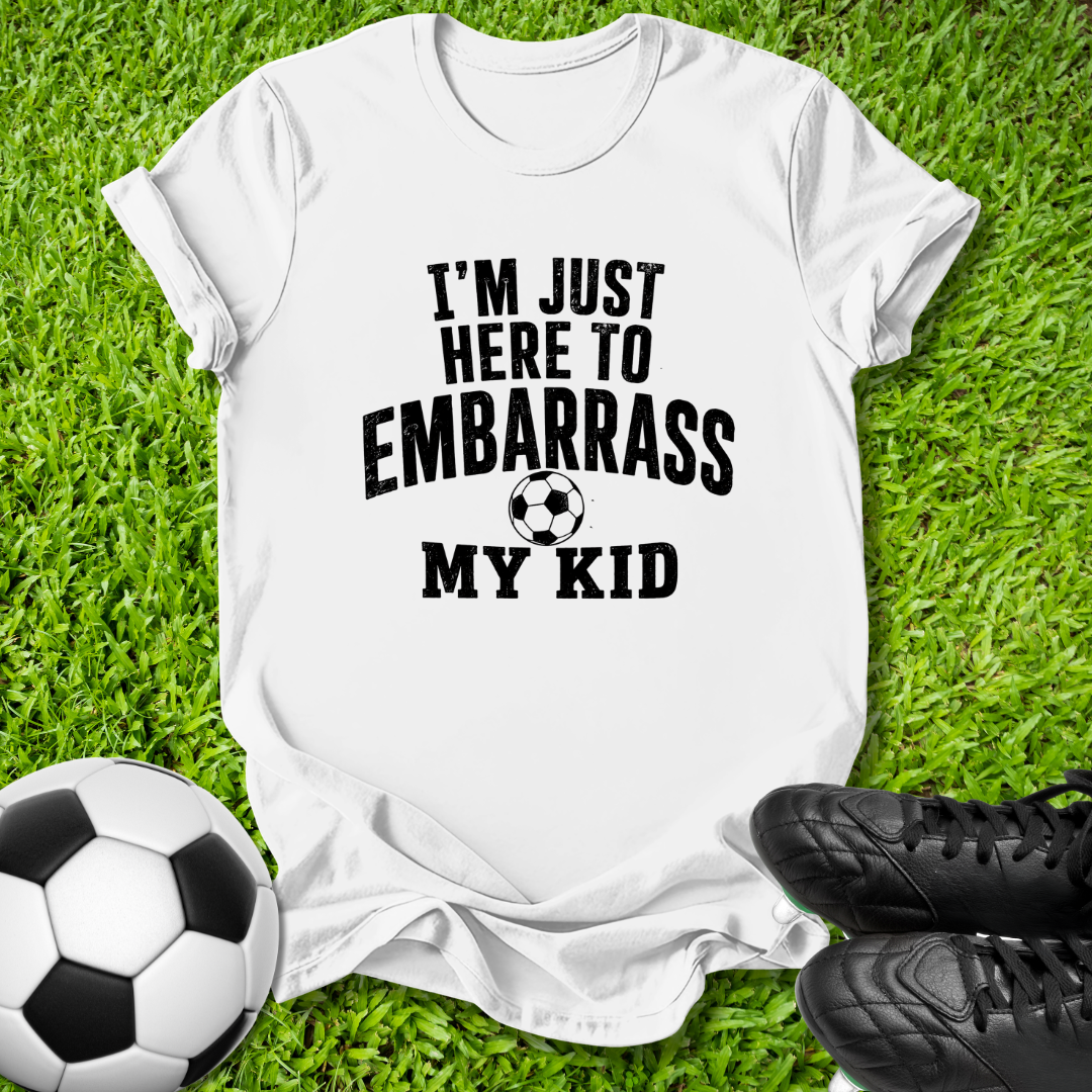 A white short-sleeve graphic T-shirt laid flat on green grass, with black text reading “I'M JUST HERE TO EMBARRASS MY KID” and a soccer ball graphic. A soccer ball and black cleats are placed around it as props.
