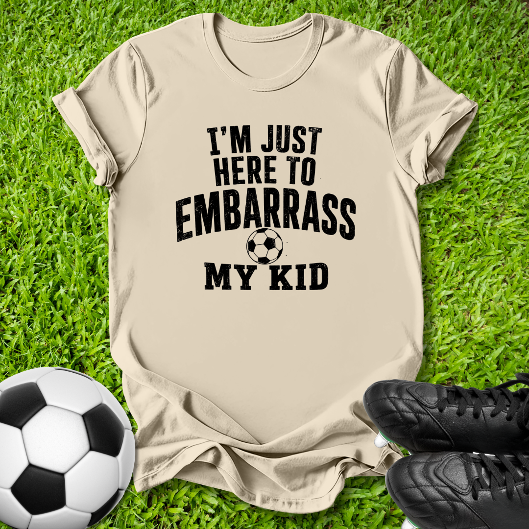 A beige graphic t-shirt laid flat on green grass, featuring the black text 'I'M JUST HERE TO EMBARRASS MY KID' with a soccer ball graphic. A soccer ball and black cleats are placed around it as props.