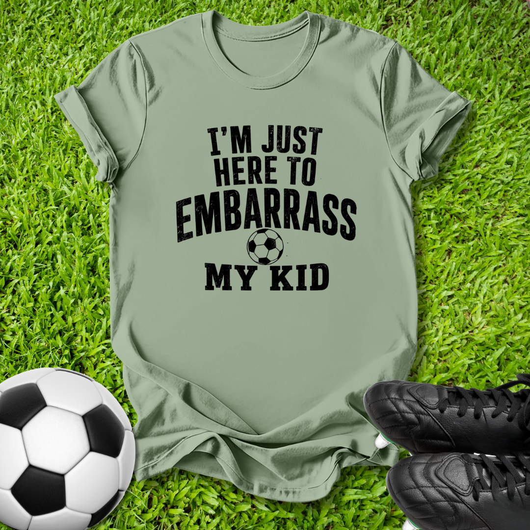 An olive green t-shirt with black text reading 'I'M JUST HERE TO EMBARRASS MY KID' and a soccer ball graphic, laid flat on green grass with a soccer ball and black cleats as props.