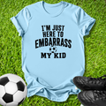 A light blue short-sleeve t-shirt with black text reading 'I'M JUST HERE TO EMBARRASS MY KID' and a soccer ball graphic, laid flat on green grass with a soccer ball and black cleats nearby.