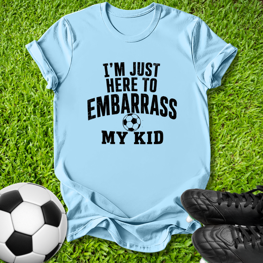 A light blue short-sleeve t-shirt with black text reading 'I'M JUST HERE TO EMBARRASS MY KID' and a soccer ball graphic, laid flat on green grass with a soccer ball and black cleats nearby.