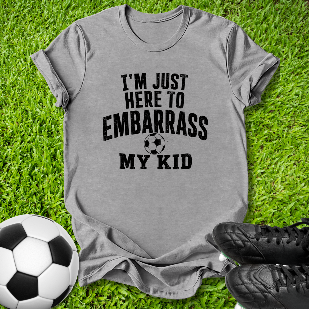 A heather gray short-sleeve t-shirt with black text reading 'I'M JUST HERE TO EMBARRASS MY KID' and a soccer ball graphic, laid flat on green grass with a soccer ball and black cleats nearby.