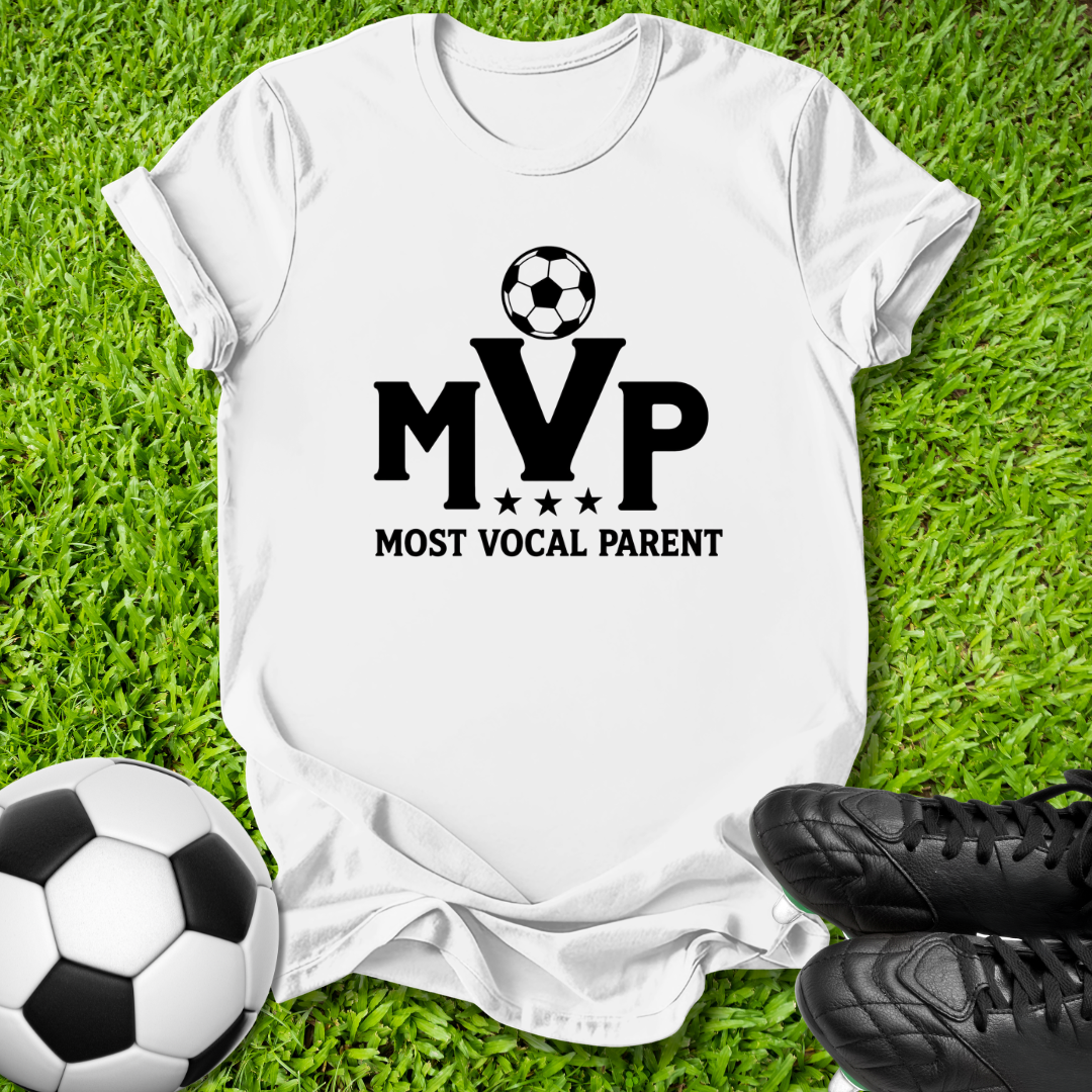 A white short-sleeve T-shirt laid flat on green grass, featuring a black soccer ball graphic above the letters 'MVP' with 'MOST VOCAL PARENT' underneath. A soccer ball and black cleats are placed around it as props.