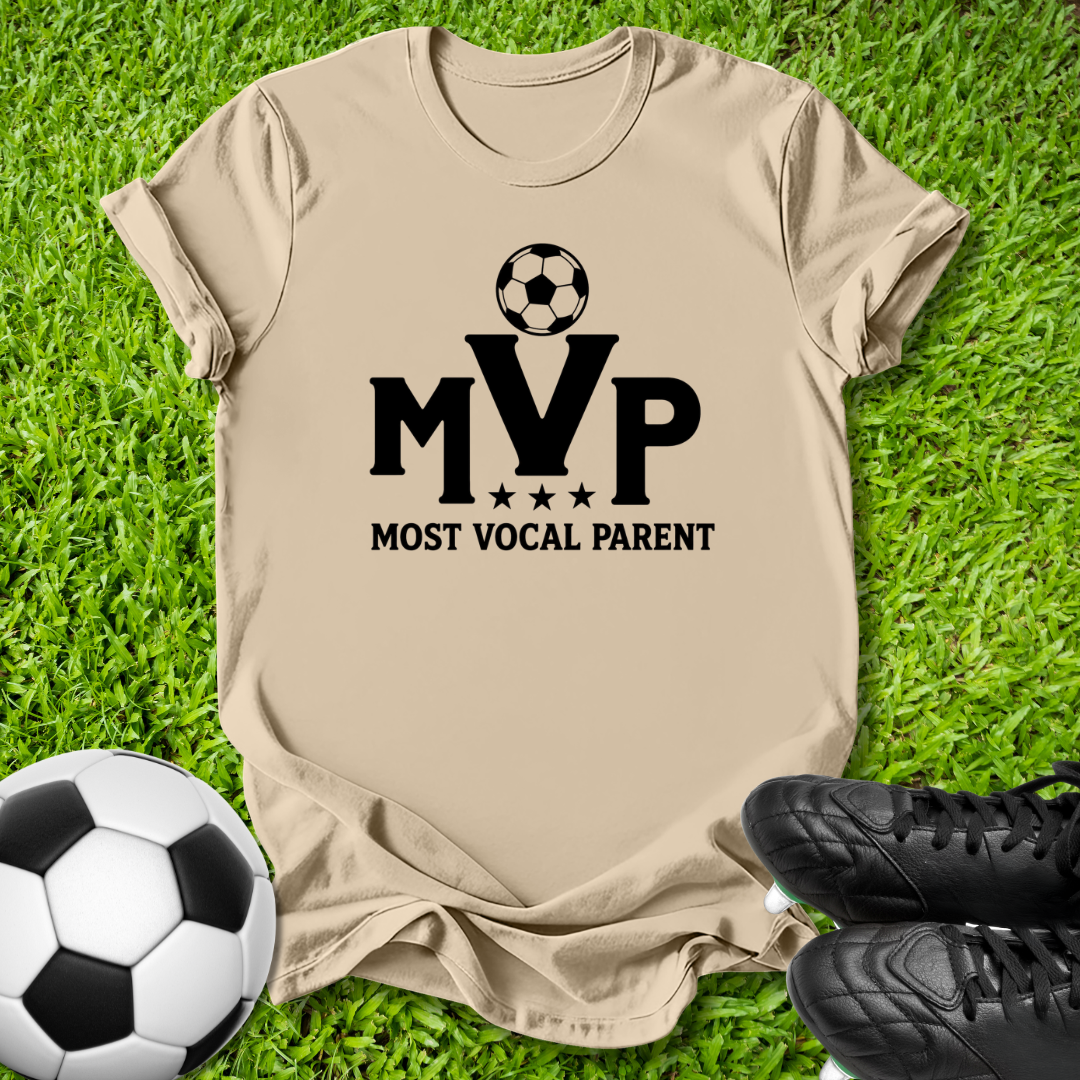 A beige short-sleeve t-shirt with a black graphic reading 'MVP MOST VOCAL PARENT' and a soccer ball icon, laid flat on green grass with a soccer ball and black cleats nearby.