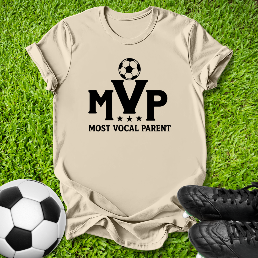 A beige short-sleeve T-shirt laid flat on green grass, featuring a black soccer ball graphic above the letters 'MVP' with 'MOST VOCAL PARENT' underneath. A soccer ball and black cleats are placed around it as props.